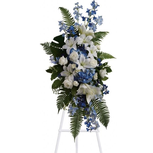 Standing funeral spray of white lilies and roses with blue hydrangea and delphinium, ferns, on a white easel.