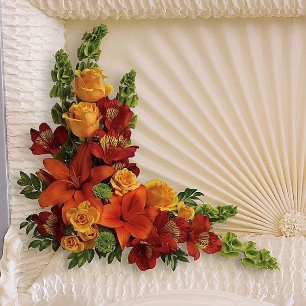 Corner casket insert with orange lilies, yellow roses, red alstroemeria, and greenery against a cream satin interior.