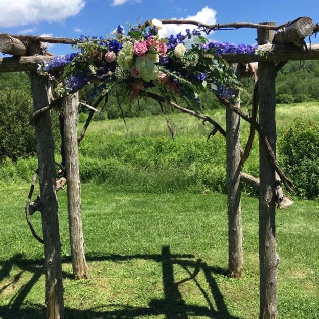 Floral wedding arch at a barn venue with roses and purple blooms beneath a blue, cloud-filled sky.