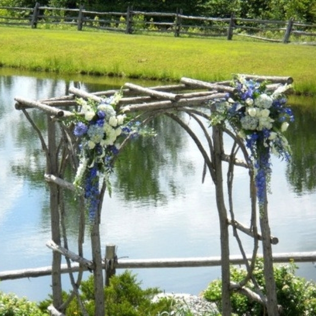 Outdoor lakeside ceremony arch made of natural wood branches adorned with white and blue flowers overlooking water.