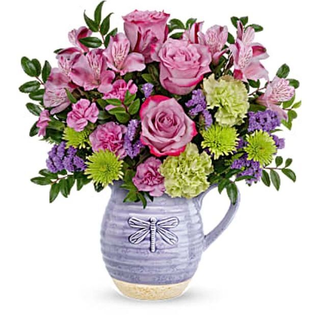 Mixed floral arrangement with pink, green, and purple blooms in a lavender pitcher featuring a raised dragonfly motif.