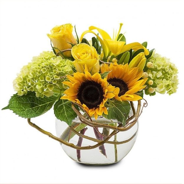 Bright floral arrangement of sunflowers, yellow roses, and green hydrangeas in a round glass vase with water.