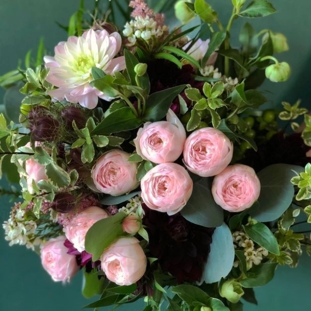 Pale pink roses and a pink dahlia nestled among burgundy blooms, small white flowers, and lush green leaves.