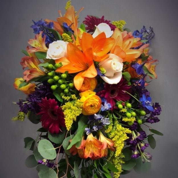Orange lilies and yellow blooms mingle with white roses, deep red daisies, purple flowers, and cascading greenery.