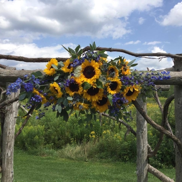 Yellow sunflowers and purple flowers decorate a rustic wooden arch overlooking a green meadow beneath a cloudy sky.