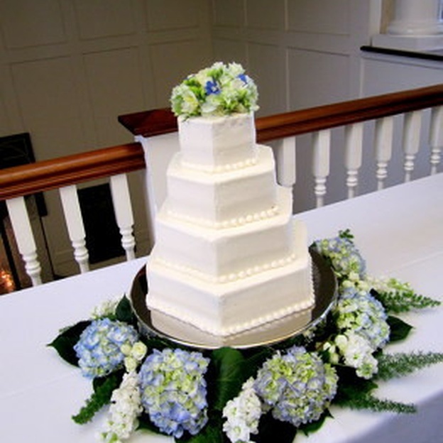 Four-tier square white wedding cake topped with flowers and surrounded by blue and pale green hydrangeas.