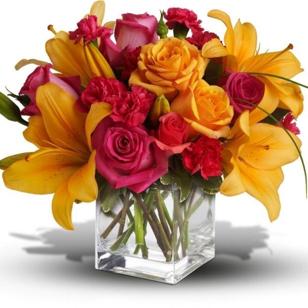 Bright mix of yellow lilies, coral and fuchsia roses, and red carnations in a clear cube vase with visible stems.