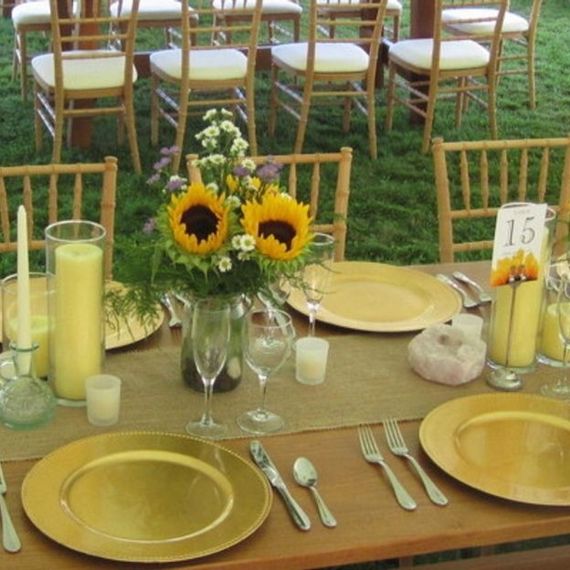 Wedding table with sunflower centerpiece, gold chargers, candles, and table number 15; chiavari chairs on grass.